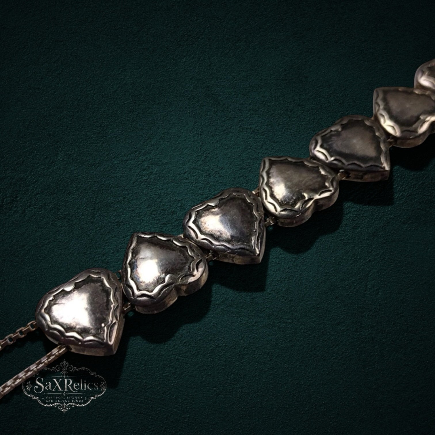 My Store SAXRelics vintage silver-tone heart link bracelet with intricate sculpted details on a dark green background.